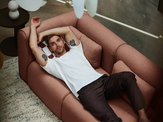 Man in white shirt lies on a brown sofa in a modern room, with a textured rug and round black side table with decorative items.