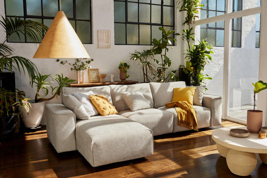 Light grey sofa with yellow cushions and throw in a sunlit room, surrounded by plants. Large pendant lamp and round coffee table nearby.
