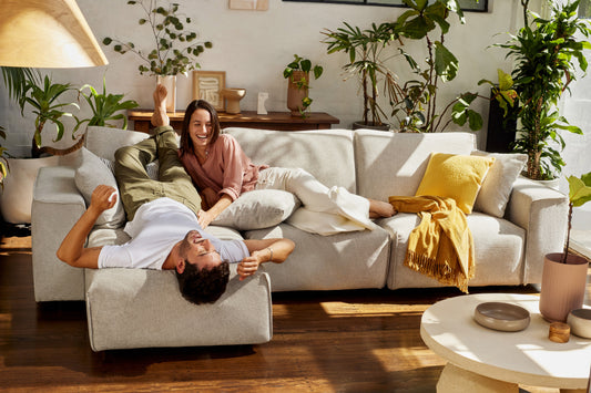 A couple relaxes on a large light grey sectional sofa surrounded by indoor plants. Sunlight streams through, highlighting a yellow cushion and throw.