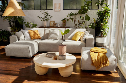 Light grey sectional sofa with mustard cushions, round beige coffee table, and indoor plants in a sunlit living room with dark wood floor.