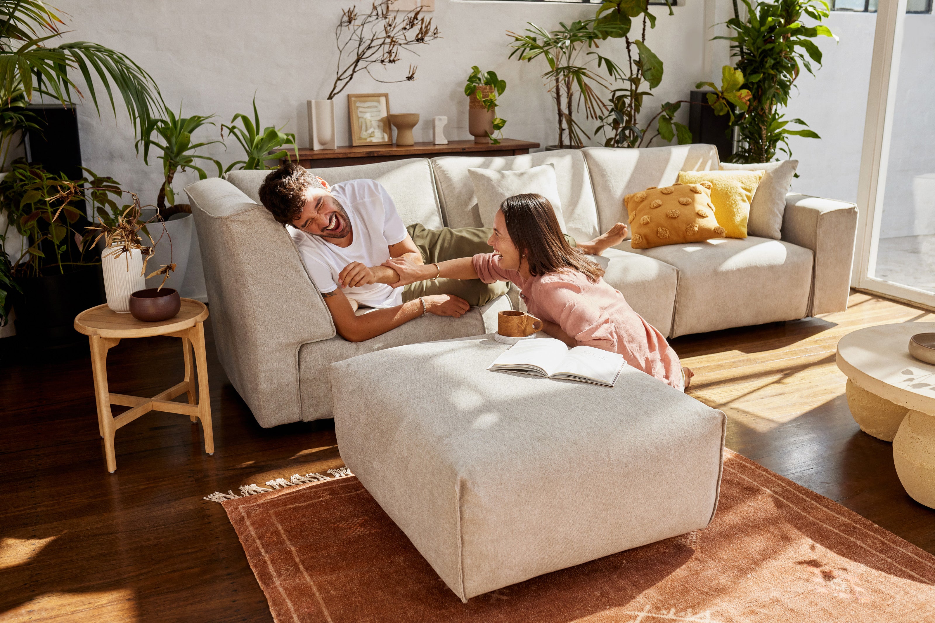 Couple laughing on a beige couch in a sunlit living room with plants, a wooden side table, and a beige ottoman on a brown rug.