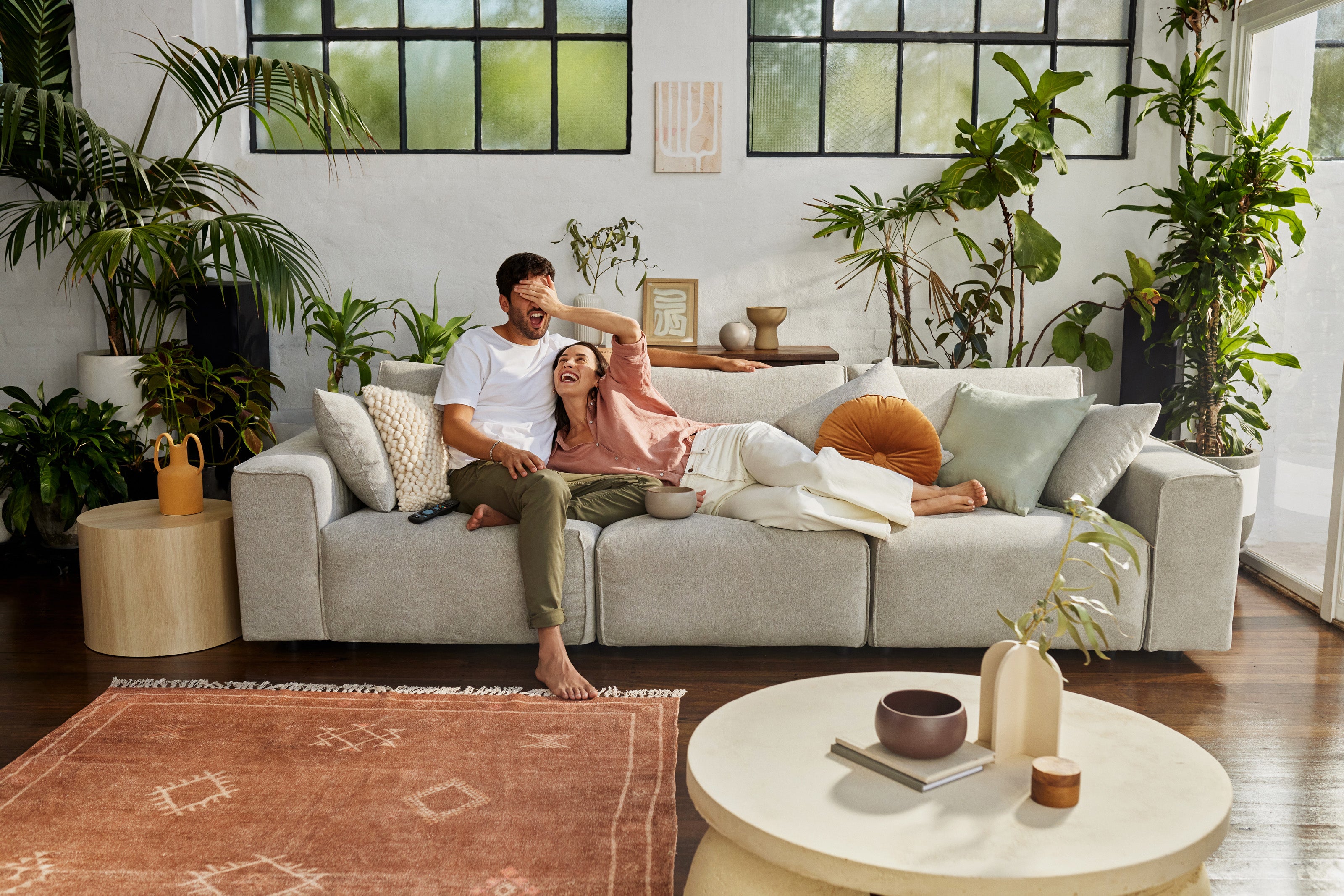 Couple relaxes on light grey sofa surrounded by indoor plants, with coffee table and rust-coloured rug in bright living room.