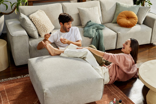 Couple laughing on light grey sectional sofa with ottoman. Wooden floor, cream and orange cushions. Nail polish bottles on rug.