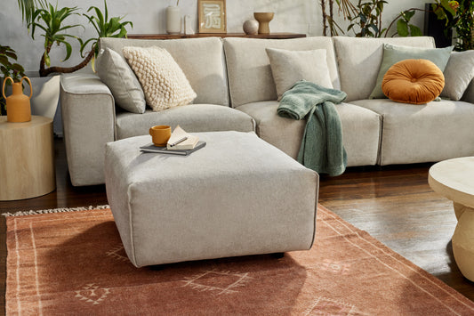 Modern living room with a light grey sofa, matching ottoman, green throw, orange and beige cushions. Wooden floors and plants accent the space.