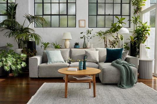 Light grey sofa with assorted cushions in a living room, surrounded by lush green plants. Wooden coffee table and white rug on floor.