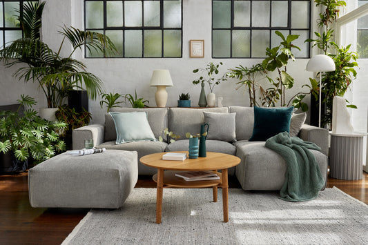 Modern living room with a light grey L-shaped sofa, wooden coffee table, green cushions, and indoor plants. Large windows and a cosy rug.