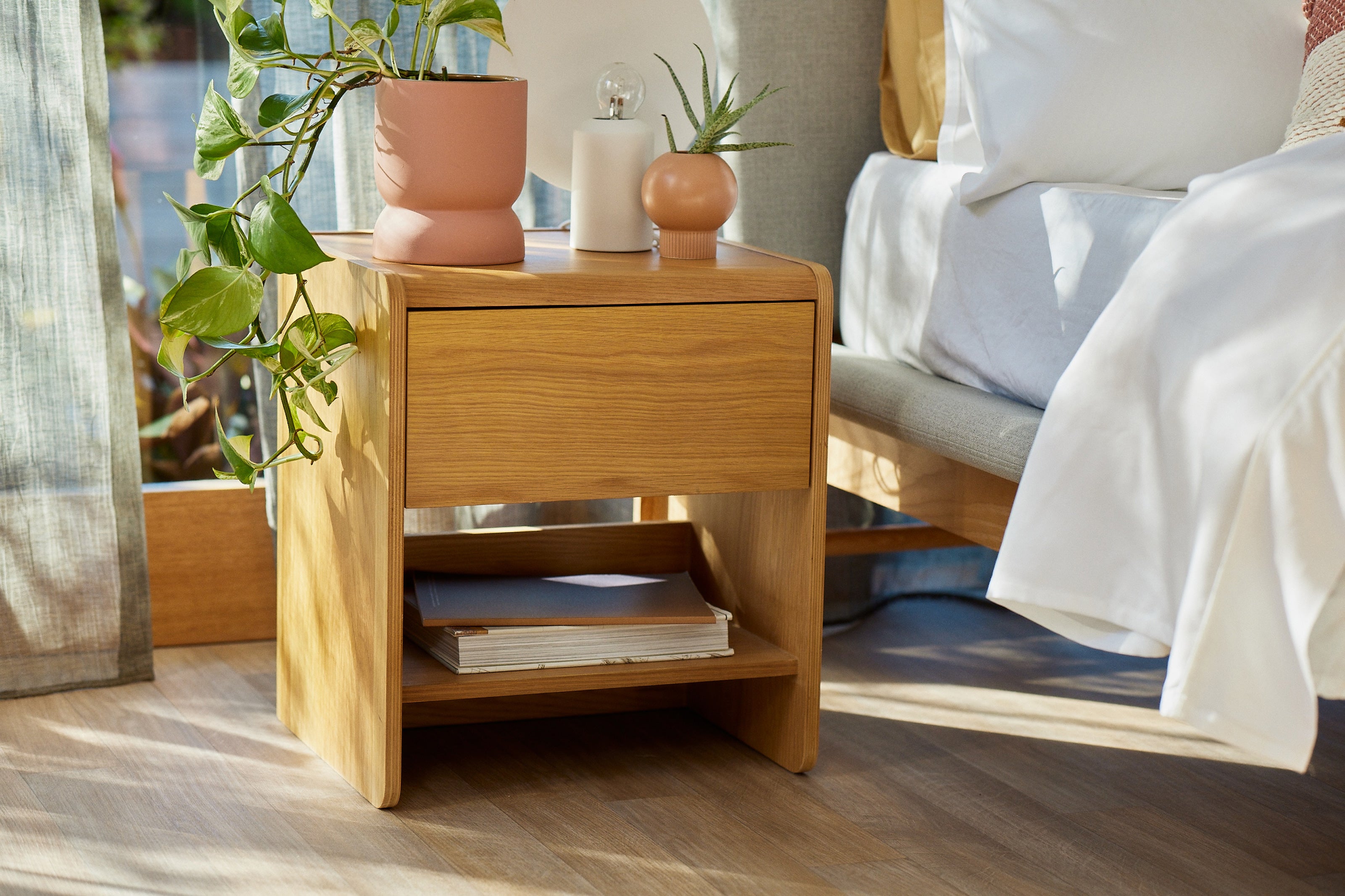 Wooden bedside table with a drawer and shelf, topped with potted plants. Next to a bed with white linens, sunlight streaming through a window.