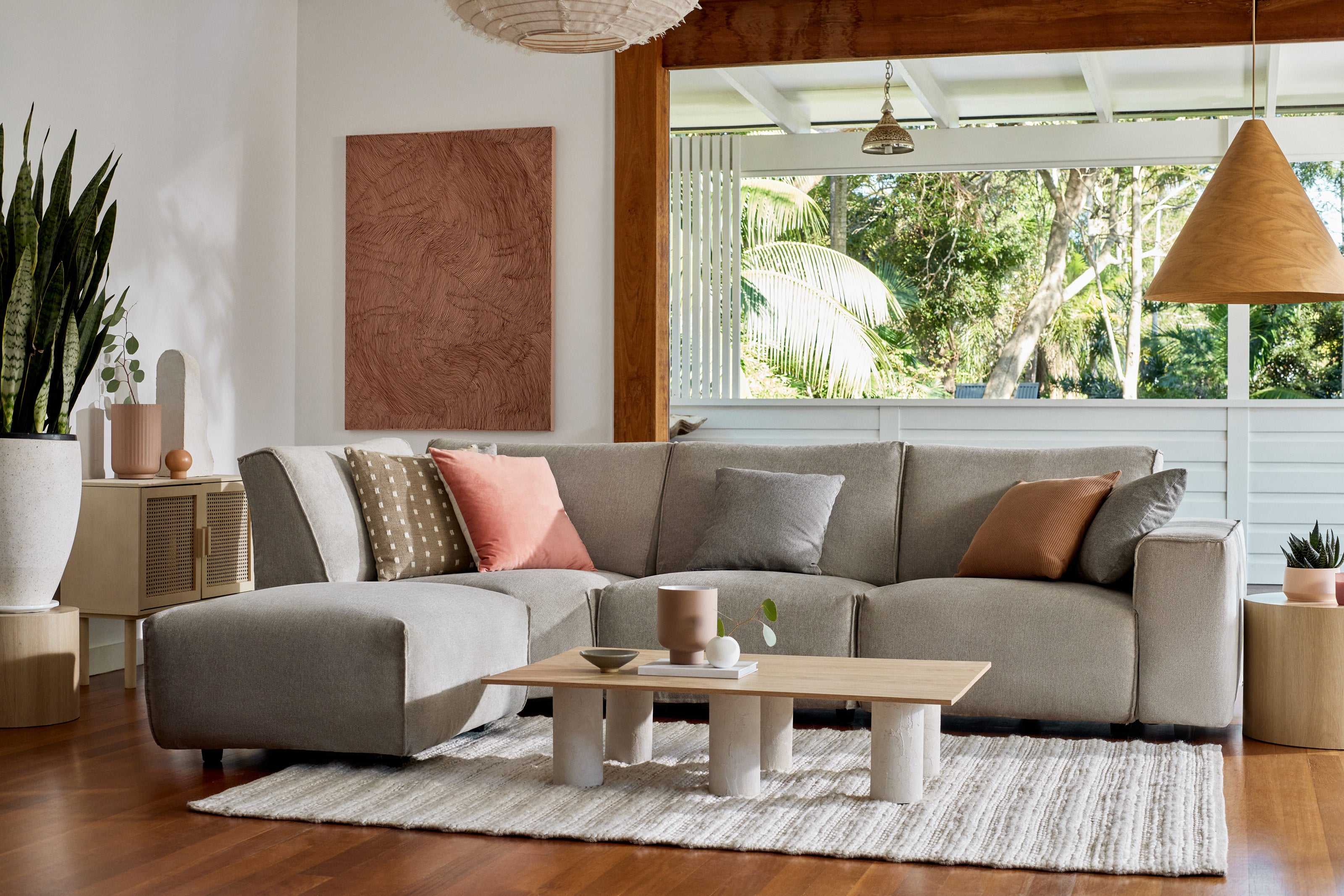 Modern living room with a light grey sectional sofa, assorted cushions in peach and grey, and a wooden coffee table on a striped rug. Large windows overlook greenery.