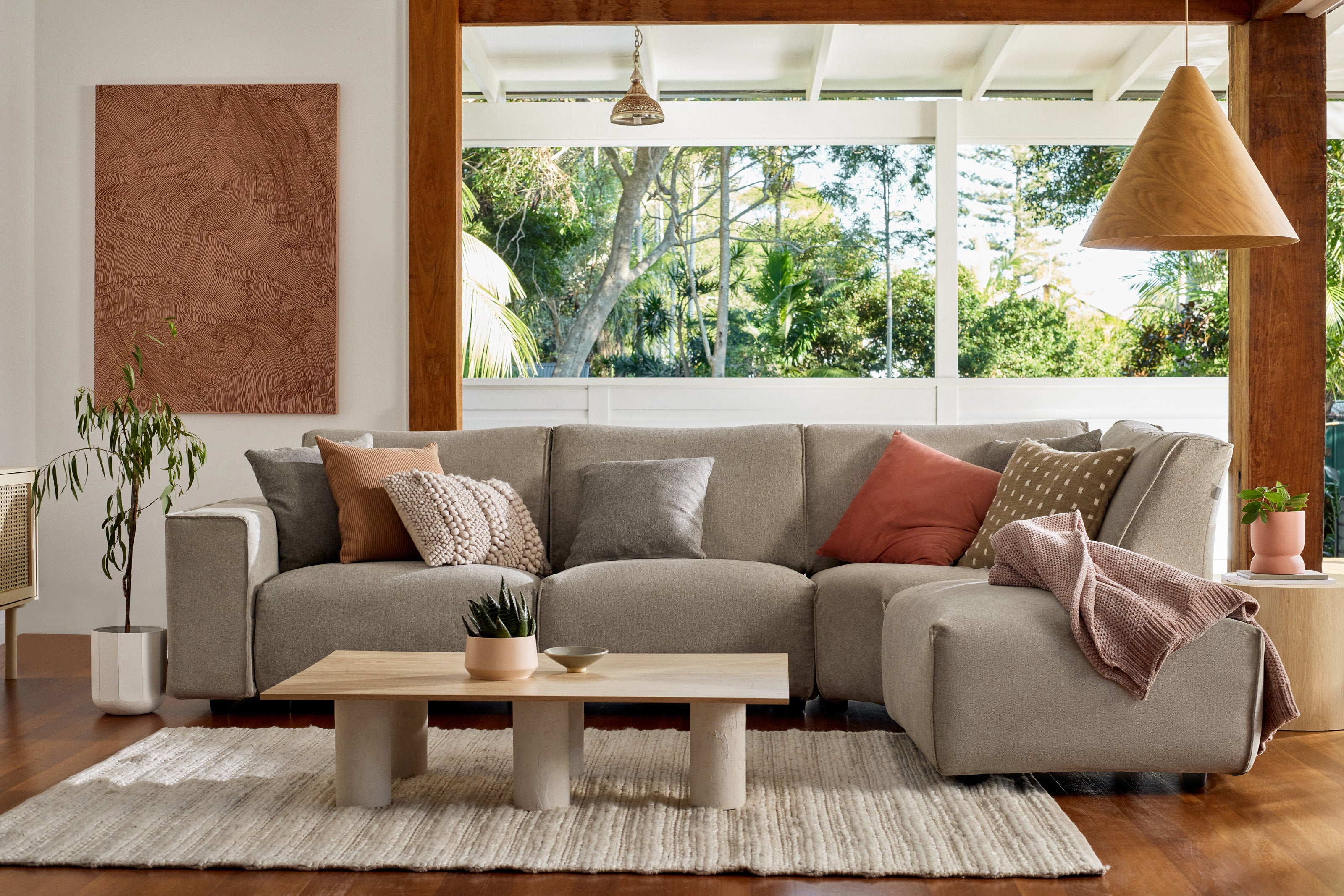 Modern living room with a large light grey sectional sofa adorned with pink and grey cushions, wooden coffee table, and floor rug. Large window view.