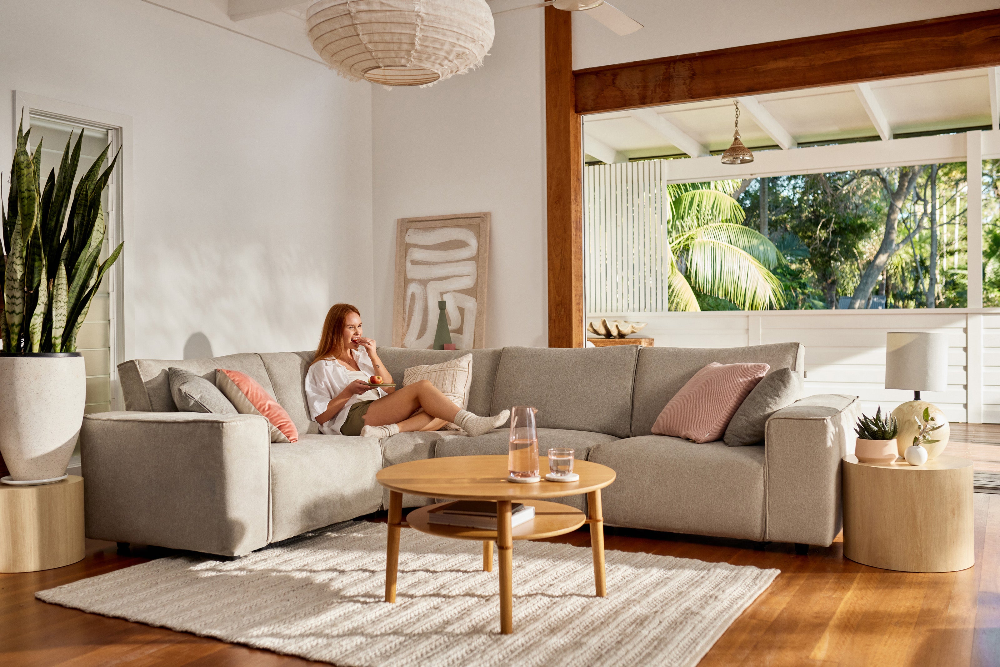 A relaxed living room with a grey sectional sofa, wooden coffee table, and large green plant, featuring a woman enjoying a book.