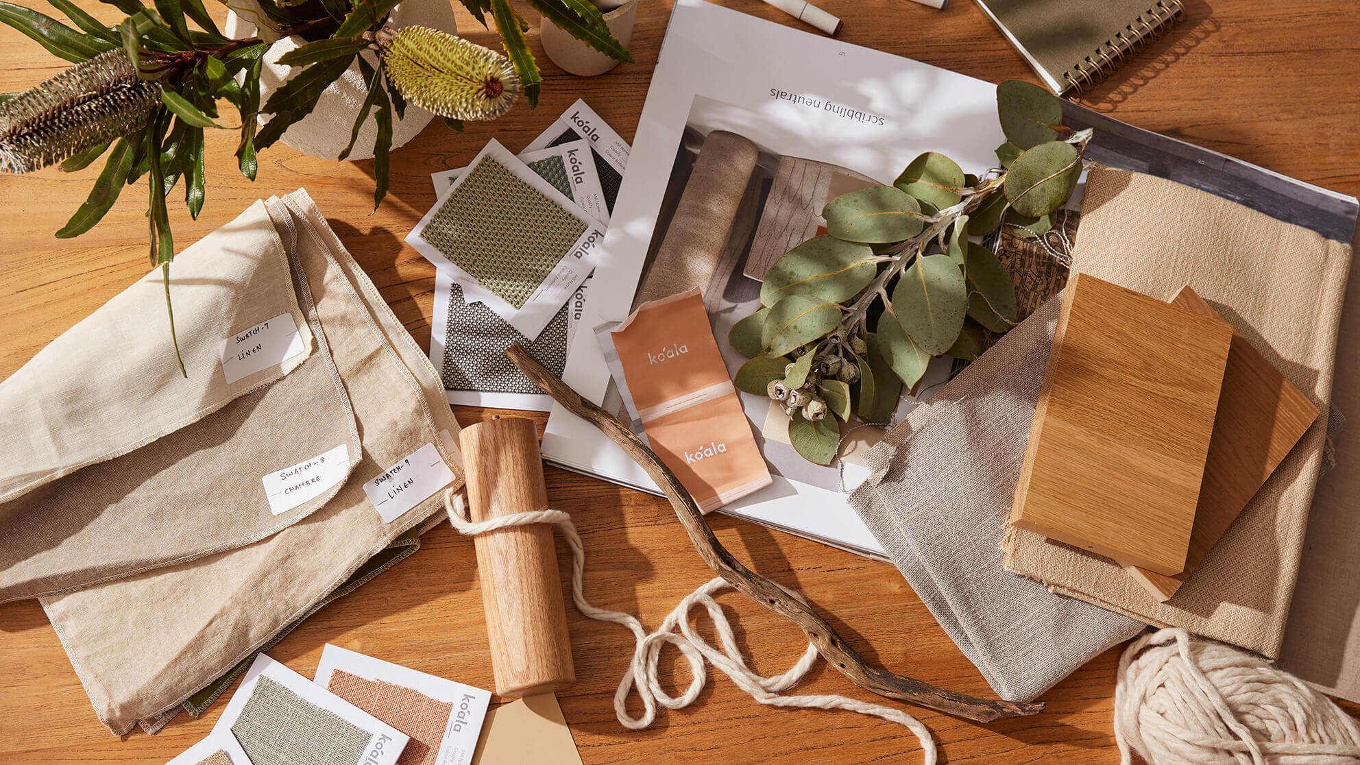 A collection of fabric swatches, natural wood samples, eucalyptus leaves, and design sketches on a wooden table under sunlight.