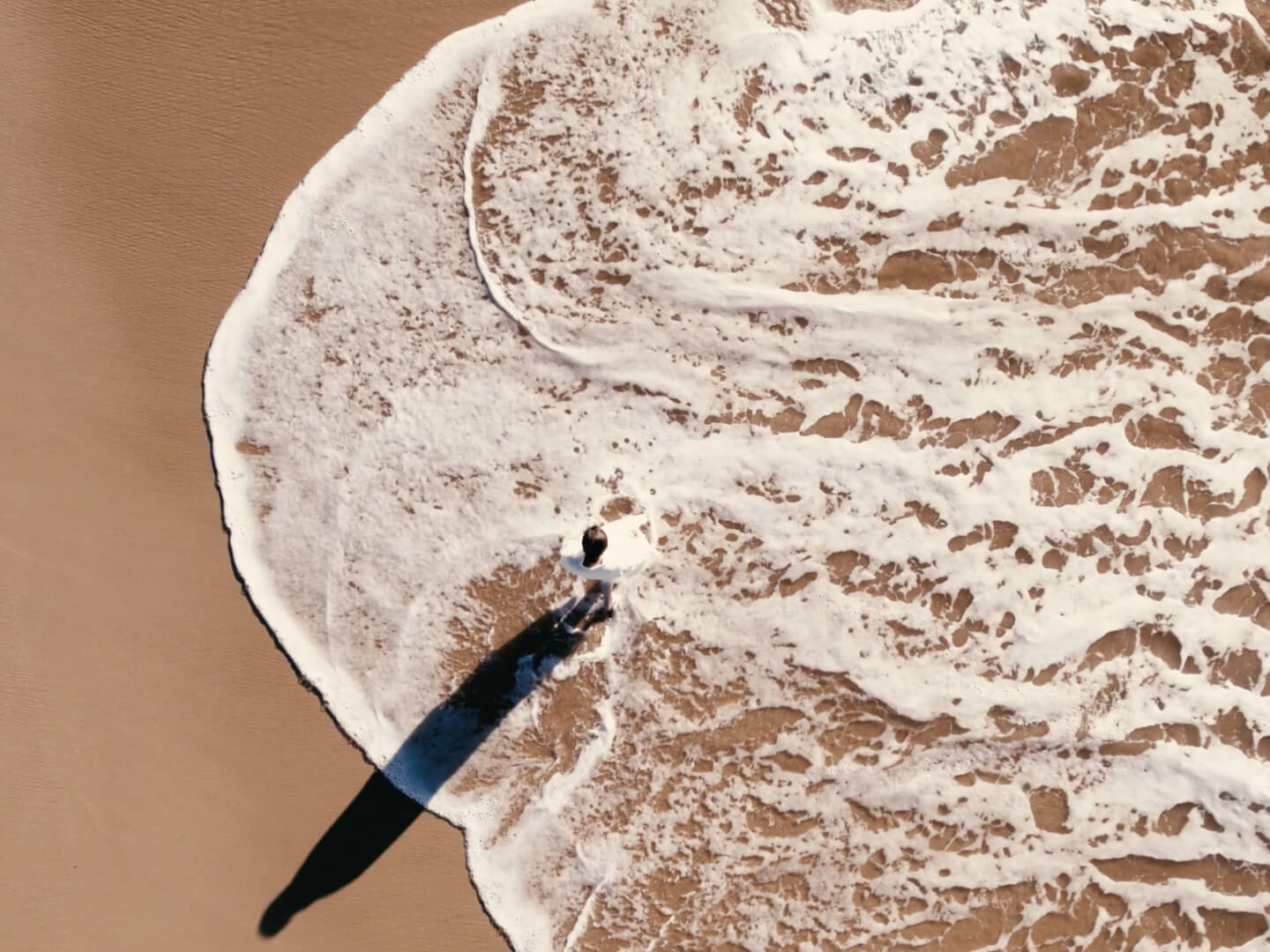 Aerial view of a person standing on a sandy beach as ocean waves cover their feet, casting a long shadow on the wet sand.