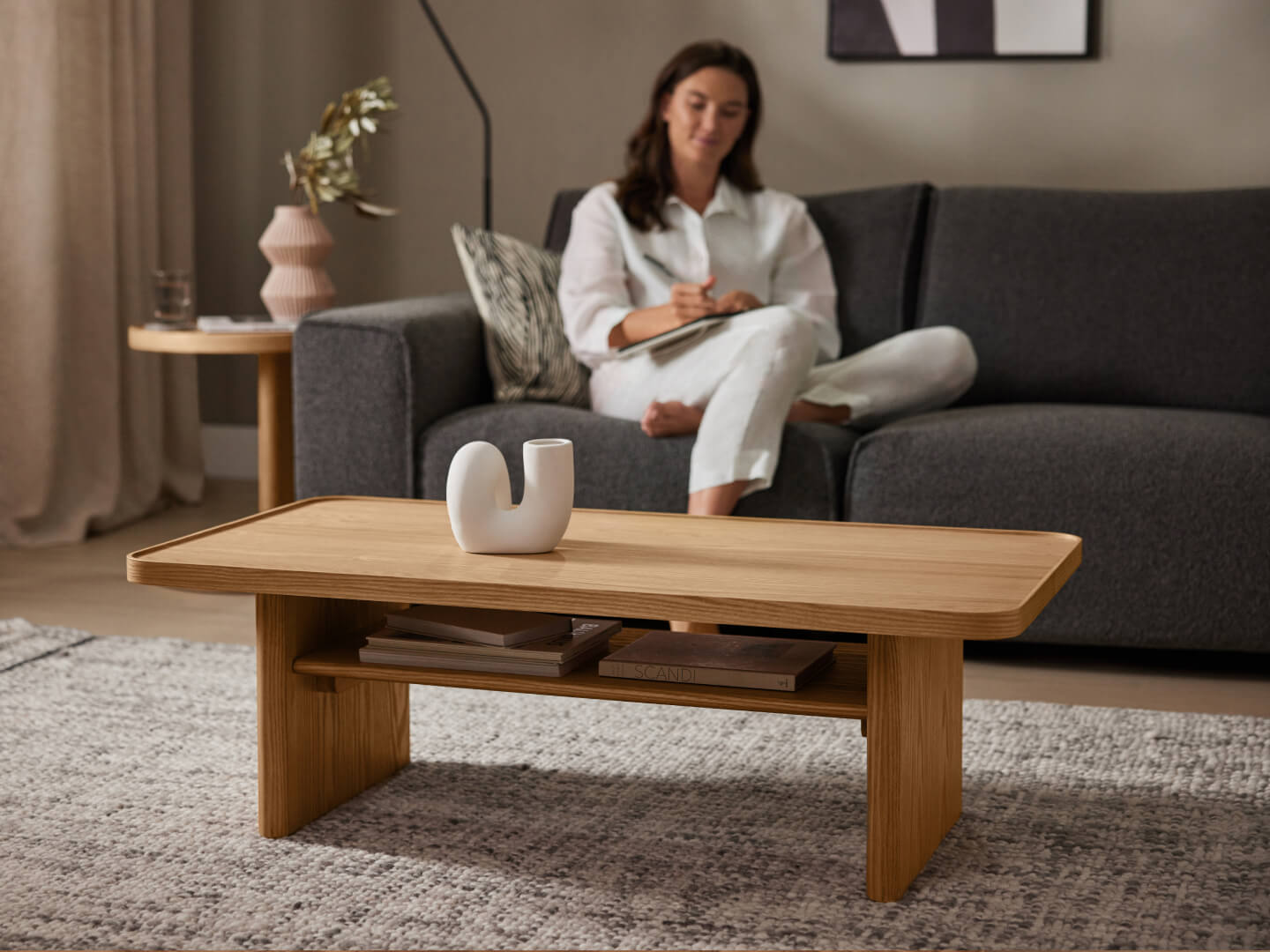 A wooden coffee table with a white sculpture on top, set on a grey rug. A person sits on a dark grey sofa in the background.