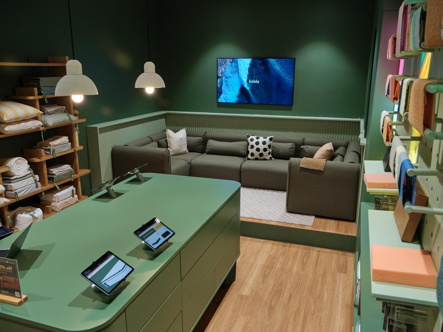 A cosy room with a dark green L-shaped couch, patterned cushions, green walls, light wood floor, pendant lights, and shelves stocked with textiles.
