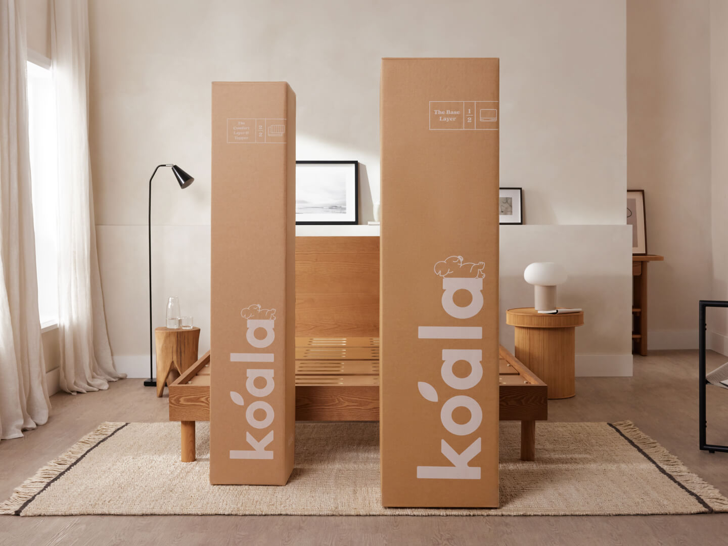 Two tall, brown cardboard boxes with "koala" branding in a minimal bedroom. Light wooden bed frame, soft lighting, and neutral decor.