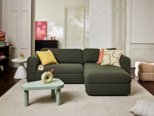 A dark green sectional sofa with yellow and beige cushions sits on a light grey rug. Nearby, a mint green coffee table holds a decorative object.