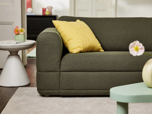 Olive green sofa with yellow cushion, white side table with flowers, on beige rug, and green coffee table in a cosy living room.