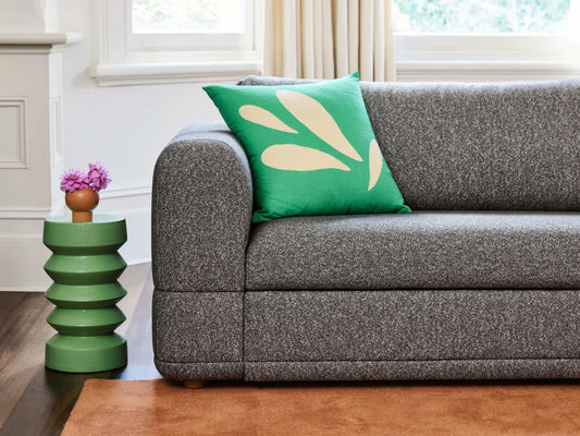 Grey couch with a green pillow featuring a beige pattern. Next to it, a green side table holds two purple flowers. Beige carpet below.