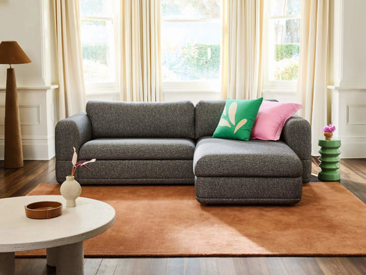 Grey sectional sofa with pink and green cushions on a brown rug, next to a round coffee table with décor; large windows in the background.