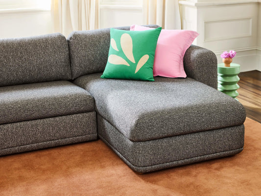 Grey corner sofa with pink and green cushions, on a brown rug in a cosy living room. White curtains and a side table with flowers.