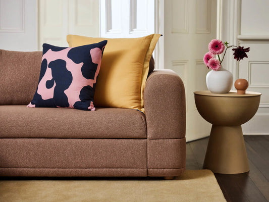 Brown sofa with yellow and pink patterned cushions next to a beige round side table. Vase with pink flowers on table. Beige rug on dark floor.
