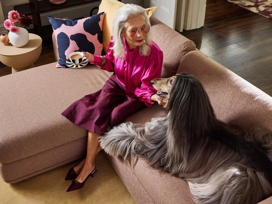 Woman in magenta blouse sitting on a beige sofa, petting a long-haired dog. Decorative pillows and a small table with flowers in the background.