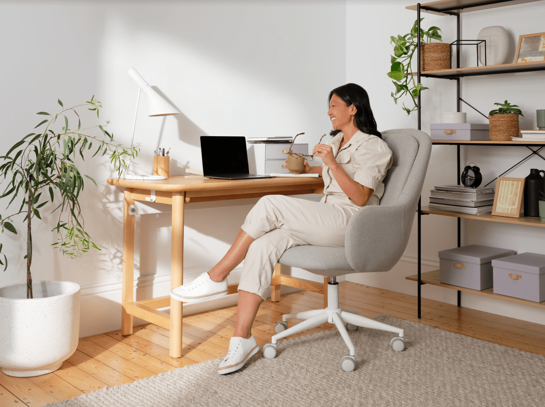 A woman sits in a light grey swivel chair at a wooden desk with a laptop, cup in hand. There's a potted plant and black shelving nearby.