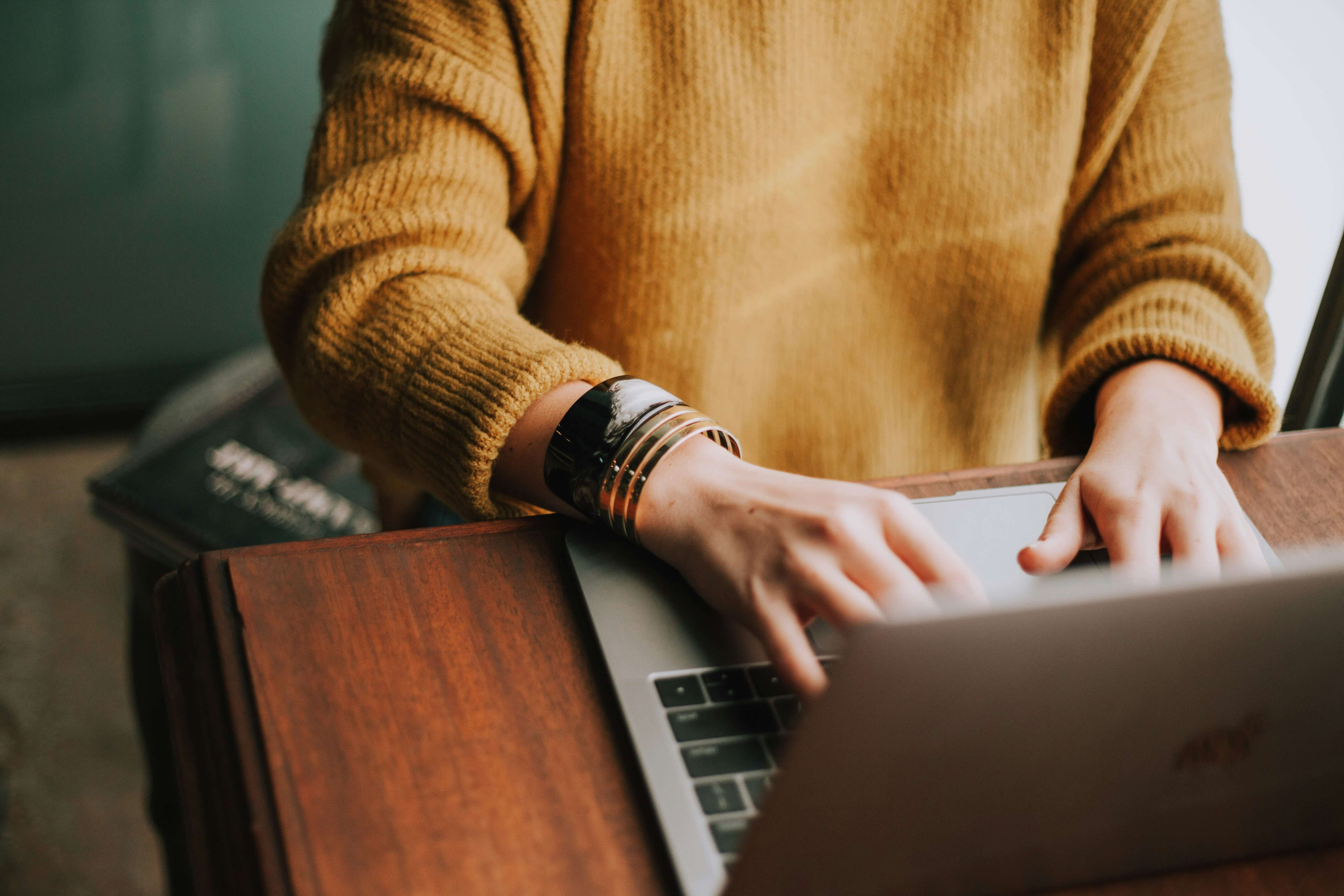 Person in a mustard jumper typing on a laptop at a brown wooden desk.