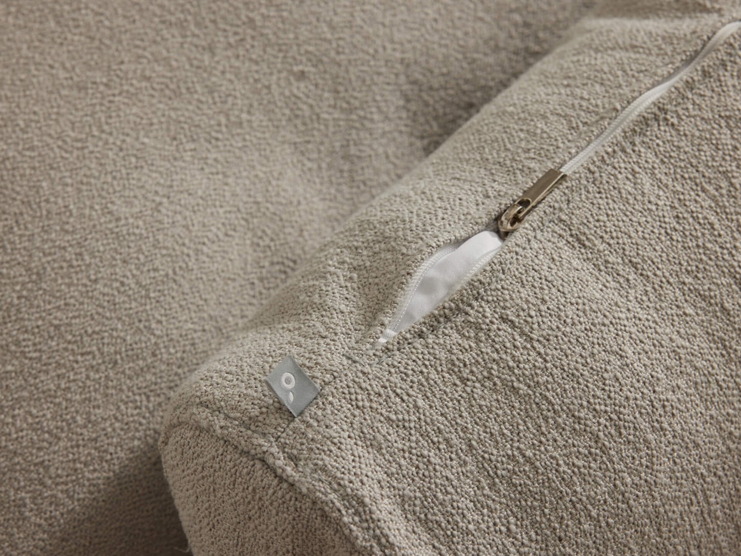 Close-up of a beige fabric cushion with a visible zip and small metal tag on a textured surface.