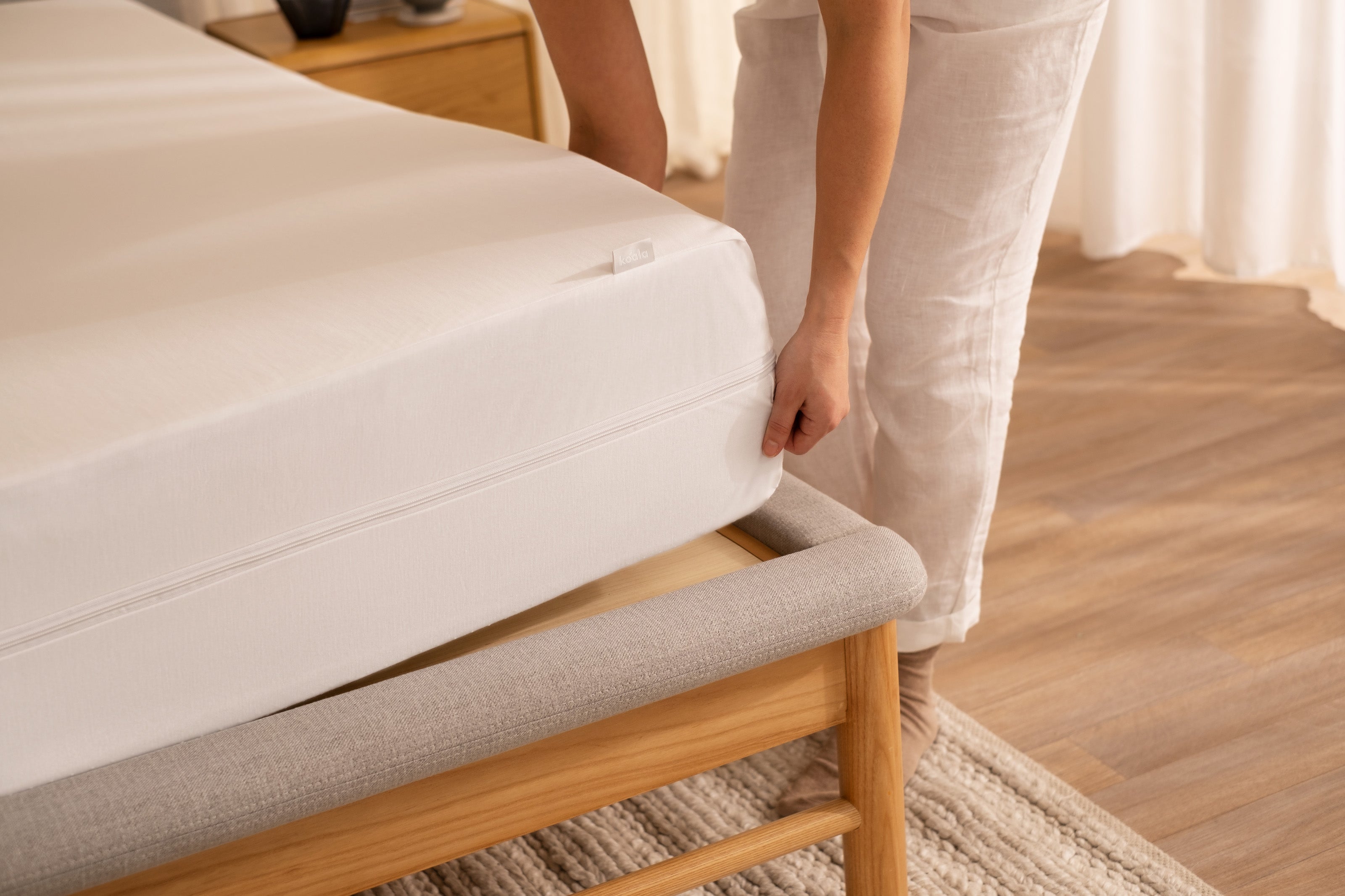 Person adjusting a white mattress on a light wooden bed frame in a bedroom with wooden flooring and a light-coloured rug.