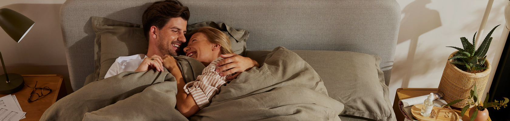 Couple laughing in bed with light grey headboard, olive green bedding, wooden bedside tables, lamp, and a potted plant.