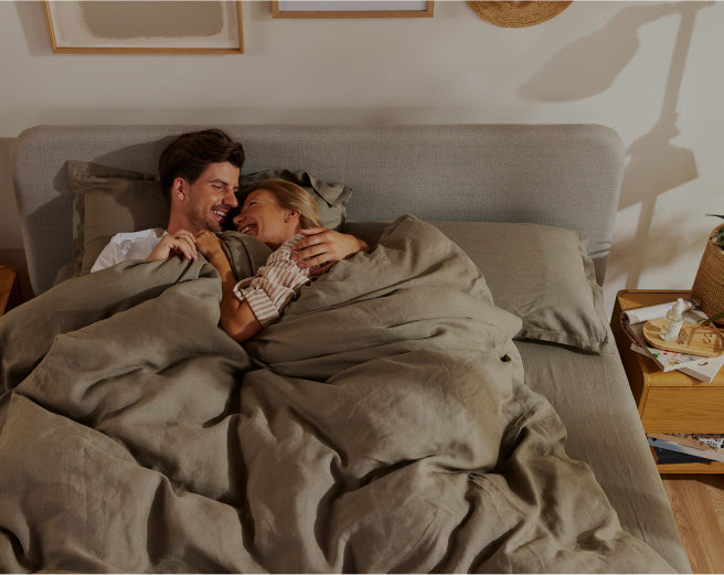 Couple smiling under olive green duvet on double bed with light grey headboard; wooden bedside tables with books and decor in a cosy bedroom setting.