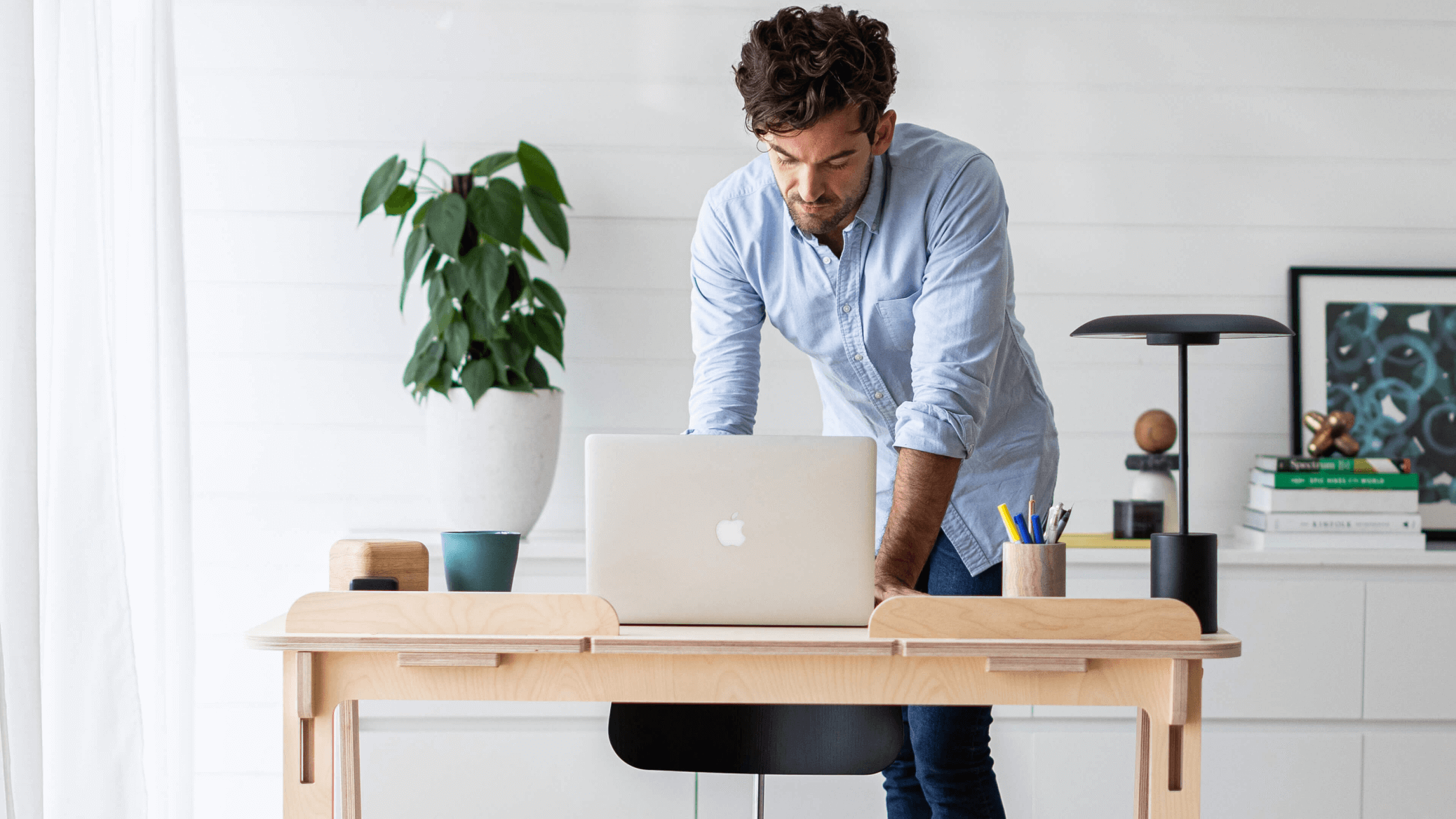 Man in a light blue shirt leans over a laptop on a light wood desk. Potted plant, books, and lamp in the background, with white walls.