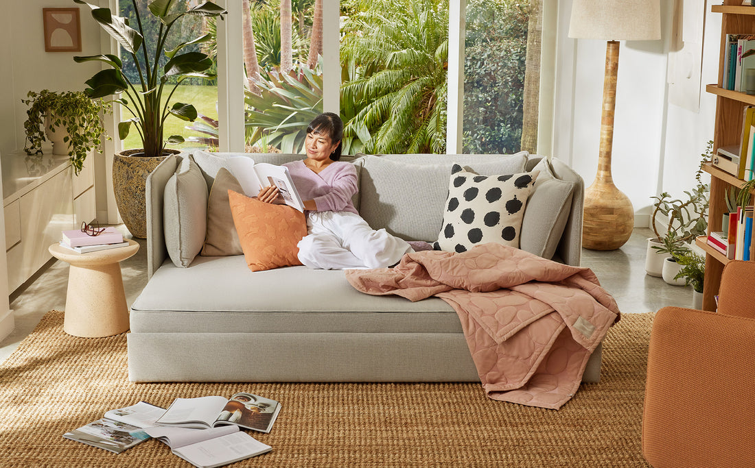 A woman relaxes on her sofa bed while reading a book in a light-filled living room with large rug