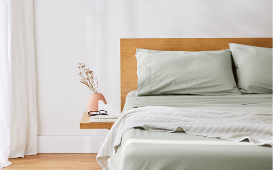 A close-up image of the corner of a bed with beautiful soft sheets, timber headboard and side table in a clean, modern bedroom