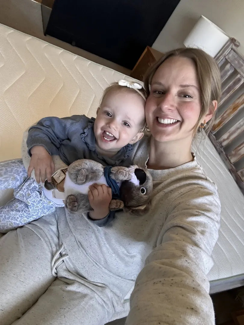 A woman and child smiling on a white mattress, with the child holding a plush toy. Brown and cream decor is visible in the background.