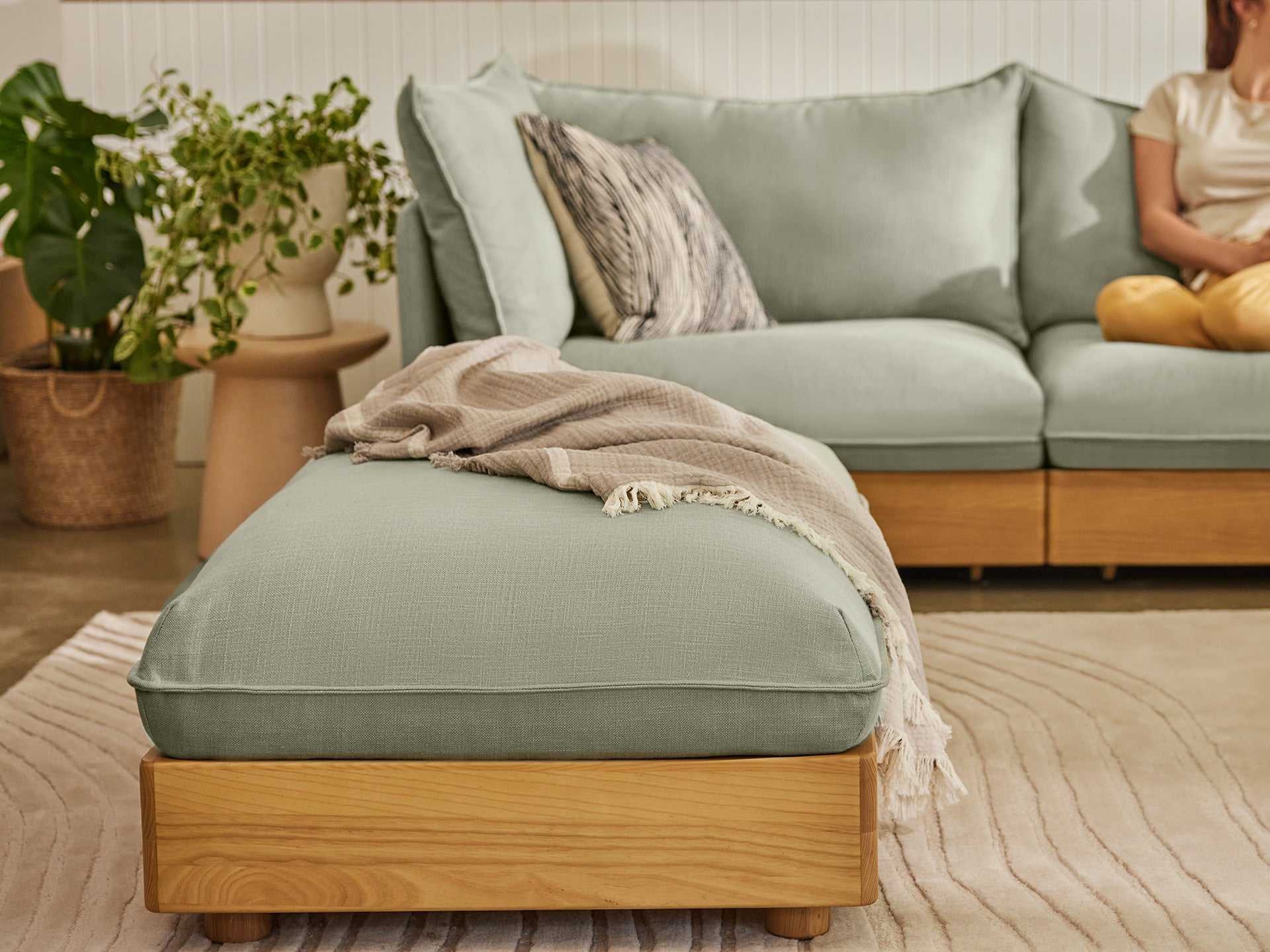 Light green sectional sofa with wooden legs, matching ottoman with draped beige throw, plant decor, and a person seated on the couch.