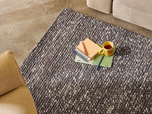 Grey textured rug with books and a yellow mug placed on top. Beige sofas surround the rug on a concrete floor.