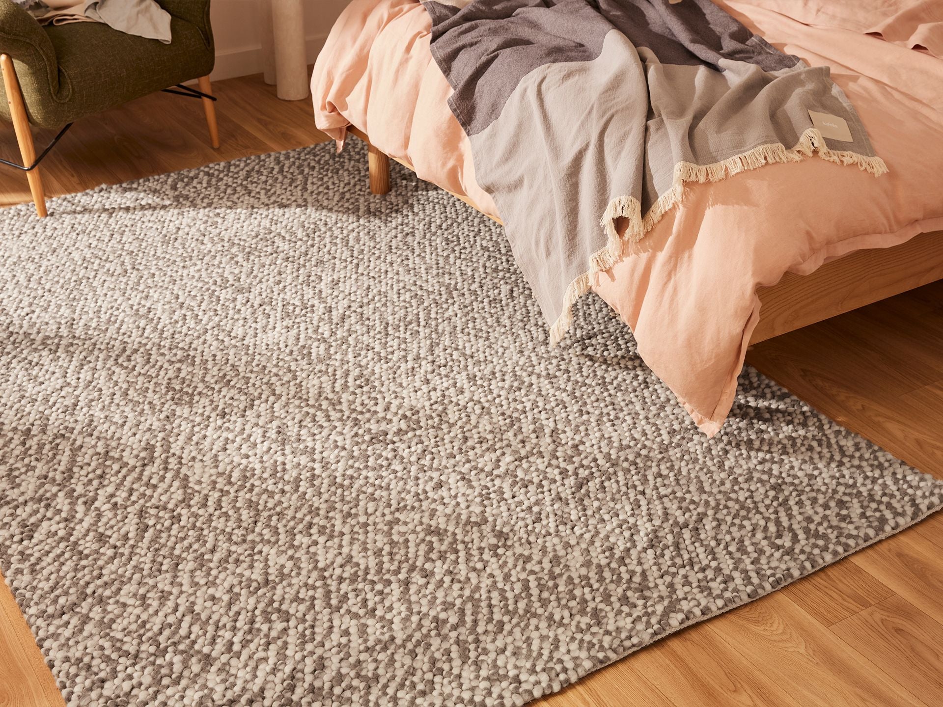 Cosy bedroom with light grey woven rug, wooden bed featuring peach and grey blankets, and an olive green chair, on wooden floorboards.