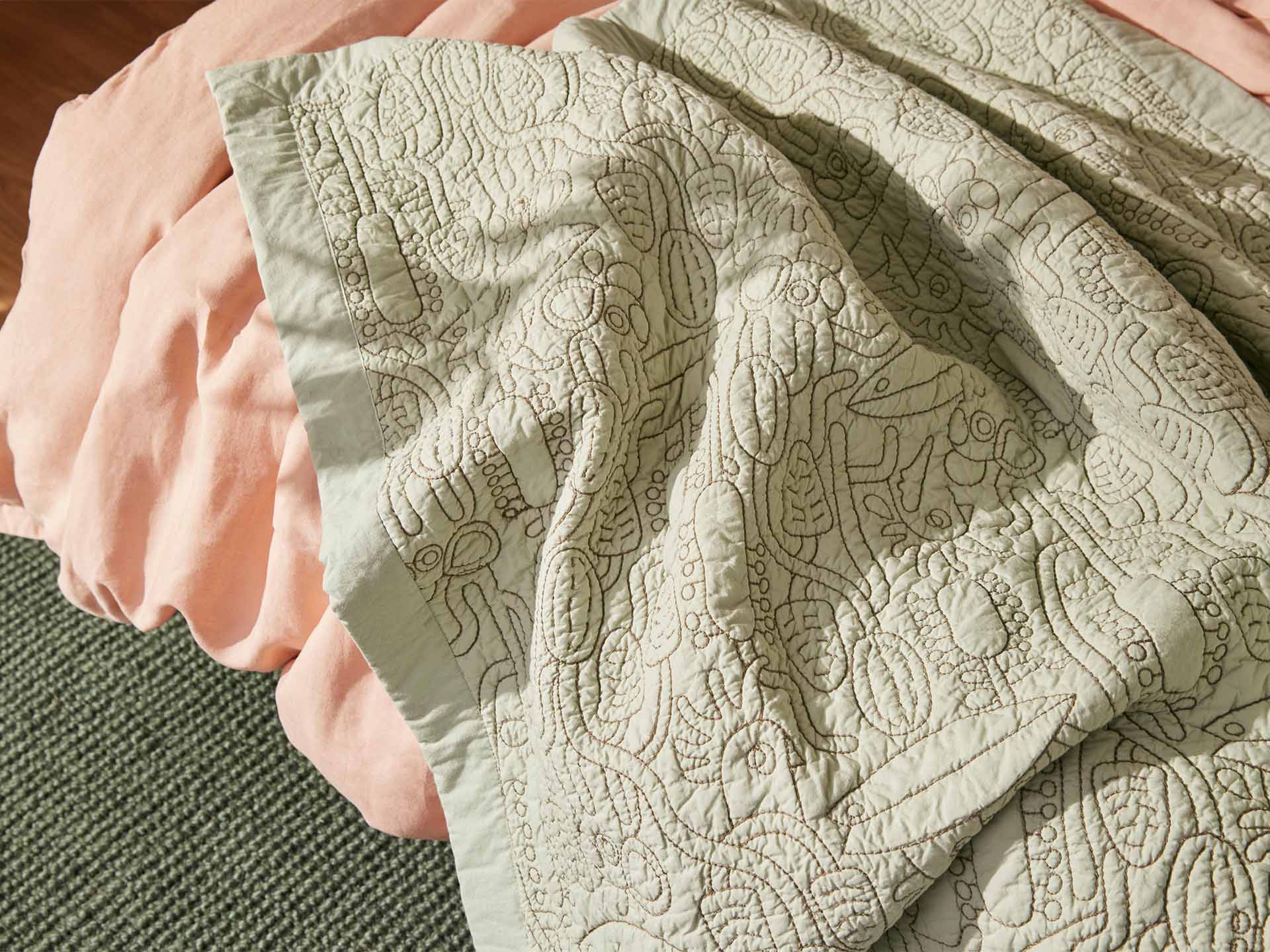 Textured quilt with intricate patterns draped over a light pink fabric on a grey carpeted floor.