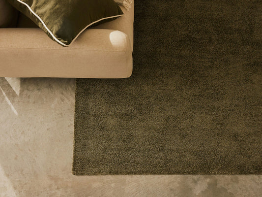 Beige couch with a green cushion next to a dark green rug on a grey concrete floor.