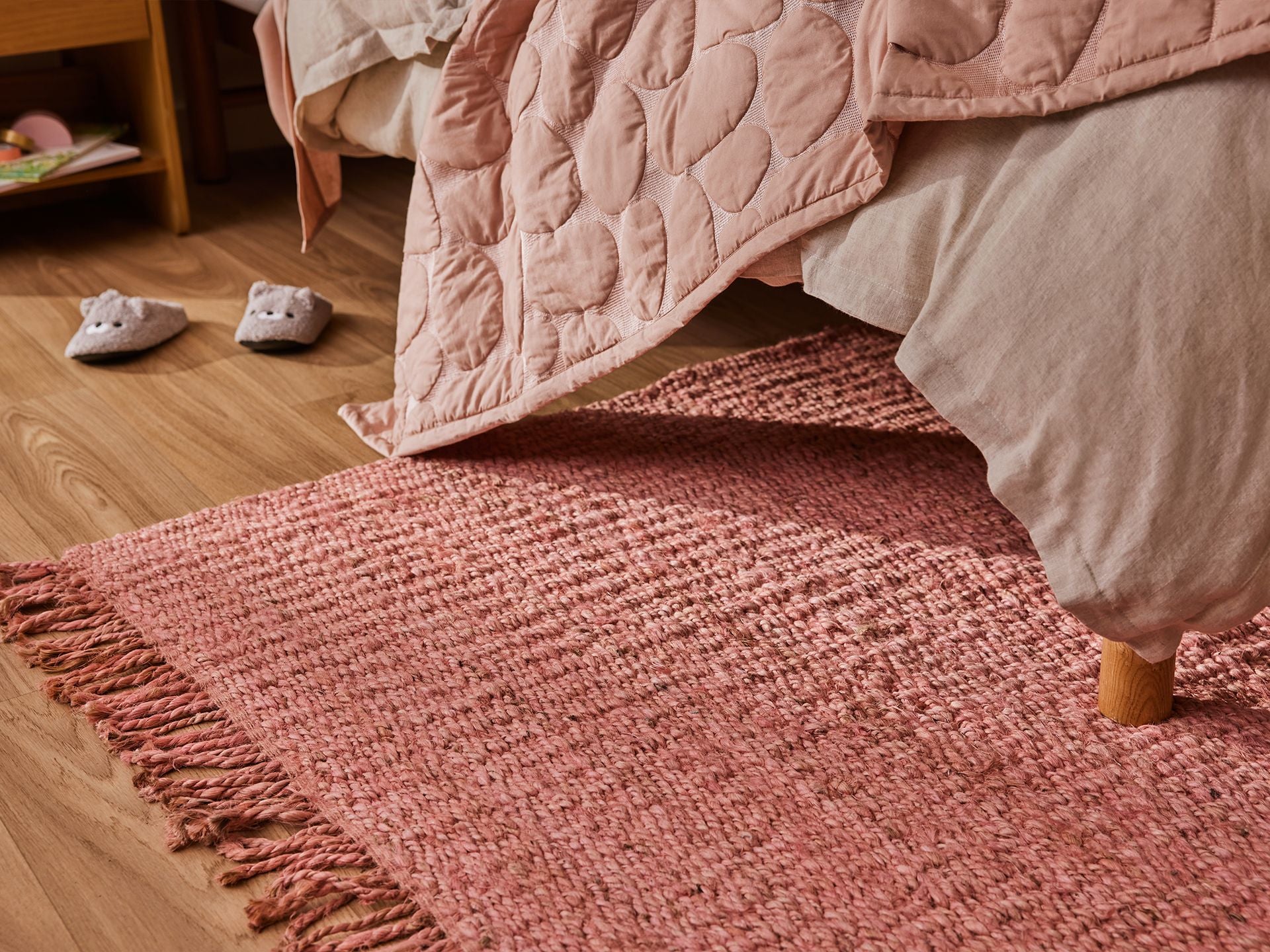 Cosy bedroom with a pink quilt and matching textured rug. Beige bedding, wooden floor, and slippers add to the warm ambience.