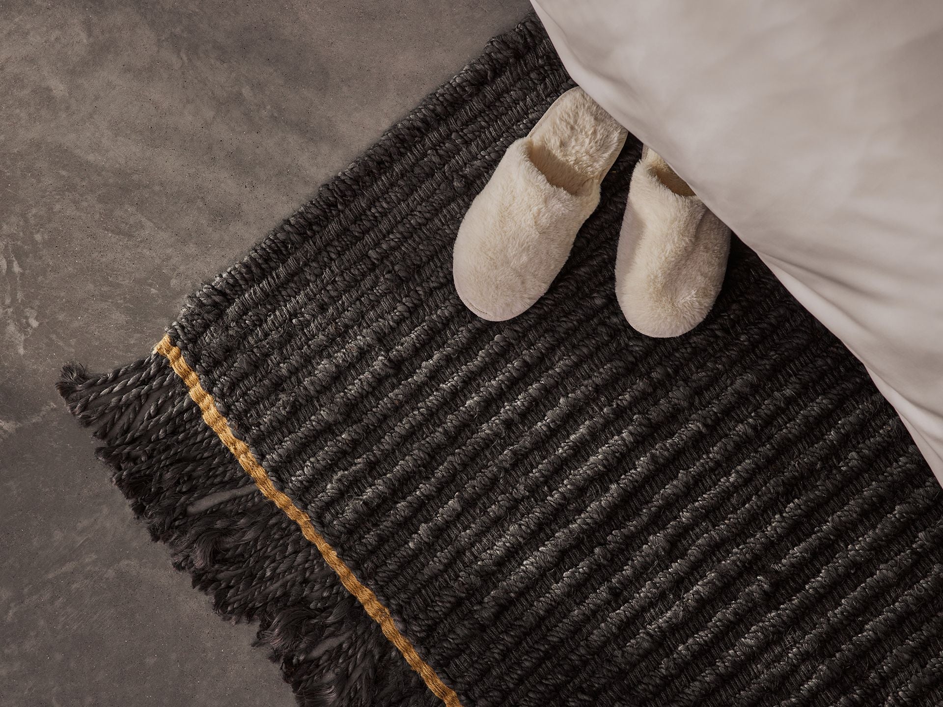 Dark grey woven rug with fringes and a gold stripe, placed beside a bed. Cream slippers rest on the rug on a grey floor.