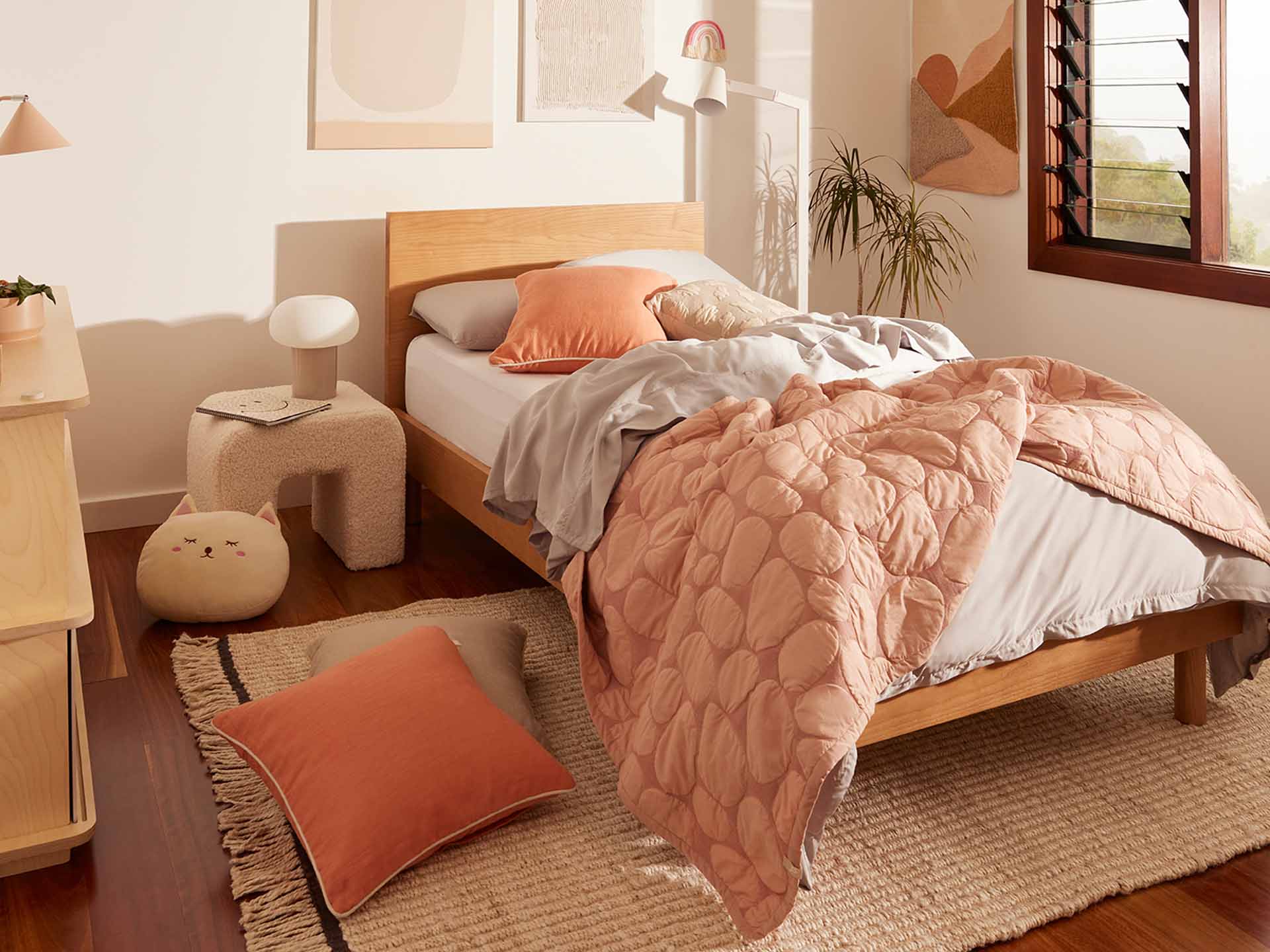 Cosy bedroom with a wooden bed, peach quilt, grey sheets, and a coral cushion. Beige armchair and lamp on a rug. Warm decor with natural light.