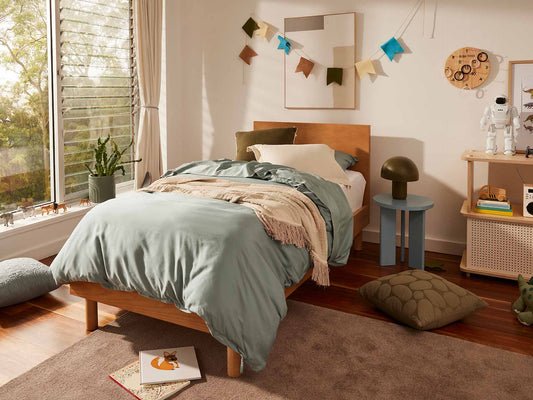 Cosy bedroom with a wooden bed, light green bedding, and decorative pillows. A lush plant and colourful garland add charm to the space.