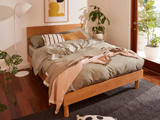 A light brown wooden bed with grey bedding, a pink throw, and decorative pillows. Room has indoor plants, a tall white lamp, and wooden floors.