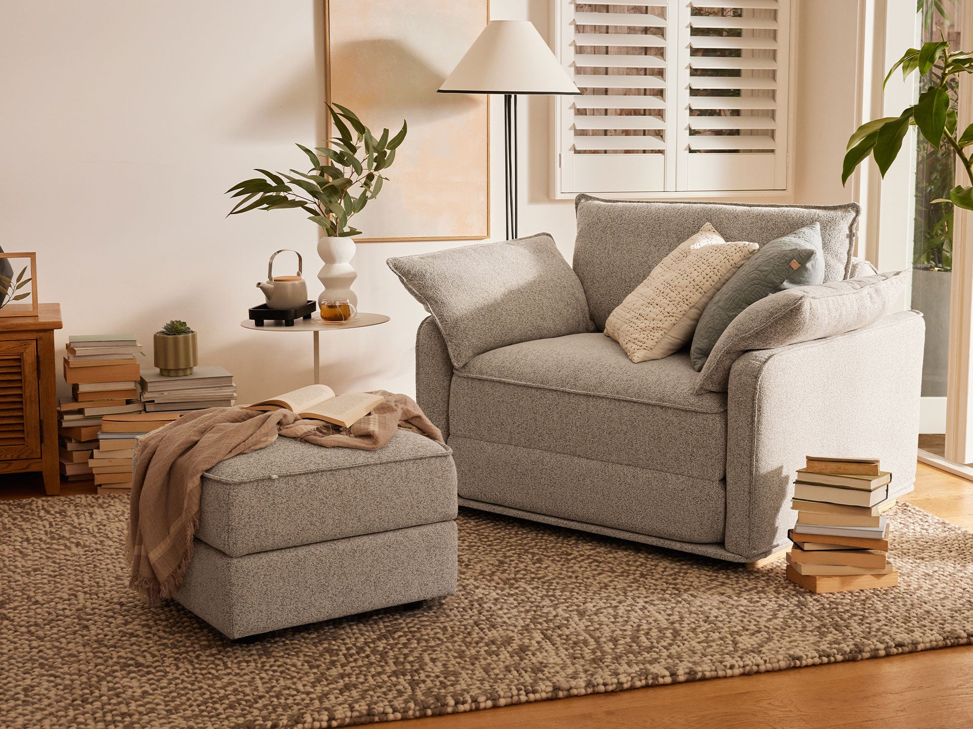 Cosy living room with grey armchair, matching ottoman, neutral cushions, and a woven rug. Stacked books and plants add warmth to the space.