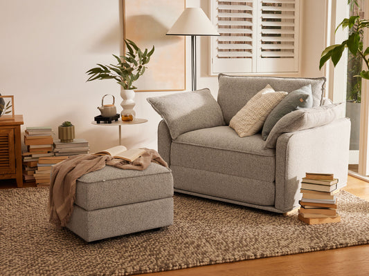 Cosy living room with grey armchair, matching ottoman, neutral cushions, and a woven rug. Stacked books and plants add warmth to the space.