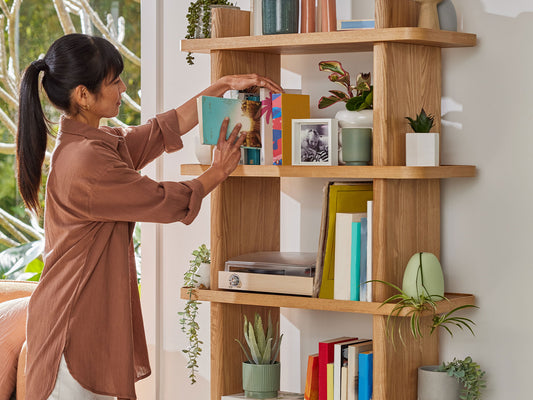 Woman arranging colourful books on a tall wooden bookshelf with various potted plants and decorative items. Light-filled room.