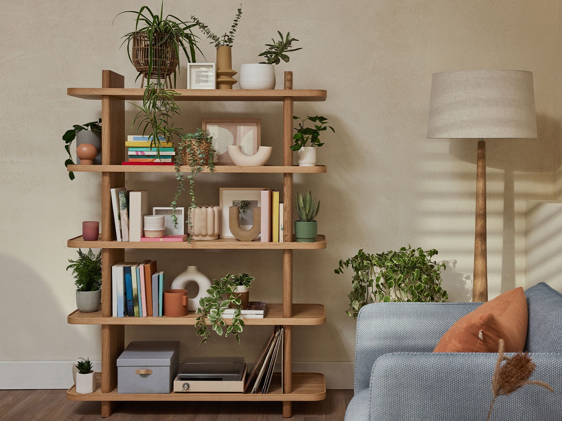 Wooden bookshelf with plants, books, and decor. Blue sofa, terracotta cushion, and beige floor lamp nearby. Modern, cosy living room setting.
